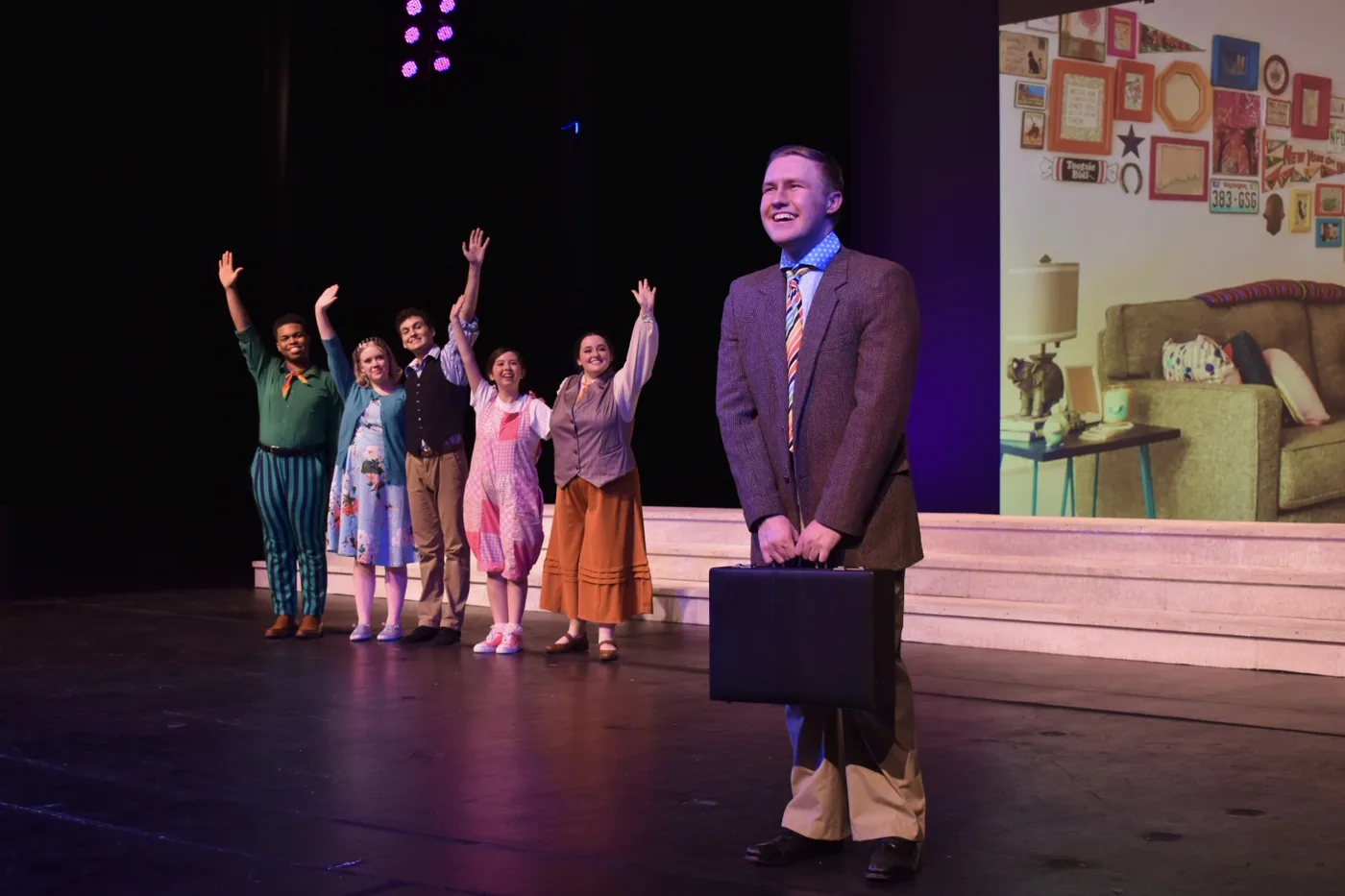 Actor downstage with briefcase, ensemble behind in a bright interior scene